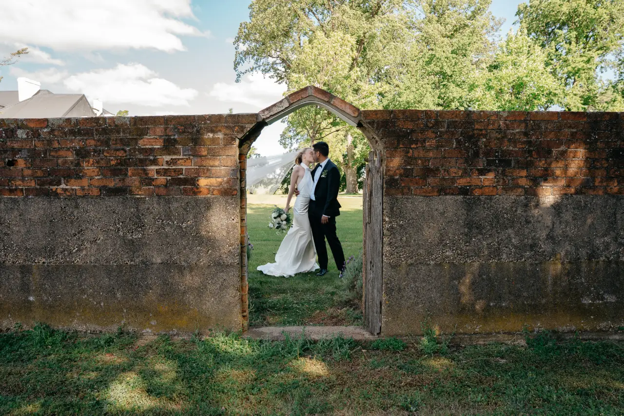A romantic, editorial-style wedding portrait of a couple sharing a kiss, beautifully framed between an elegant archway. The architectural details create a timeless, glamorous feel, capturing an intimate moment with a touch of cinematic elegance.